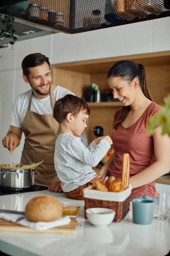 A happy family cooking together in a bright modern kitchen — a father stirring pasta, a mother smiling, and a young son on the counter.